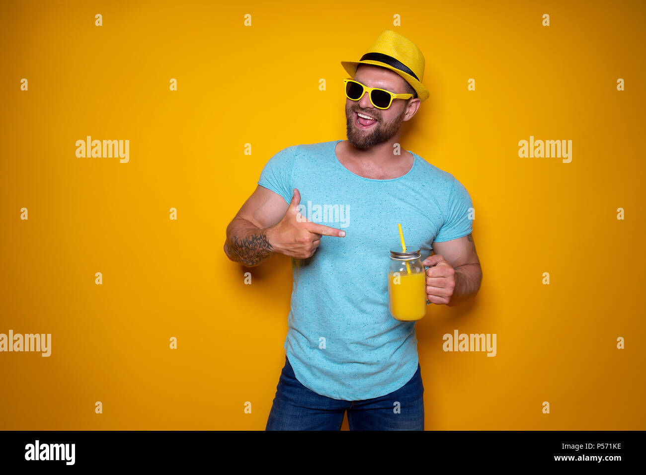 Kreative Mann in Sonnenbrille und Sommer Kleidung in gelben und blauen Farben ständigen entspannt, Zeigefinger und kreative frischen Cocktail Flasche mit Griff und auf gelben studio Hintergrund Stroh halten Stockfoto