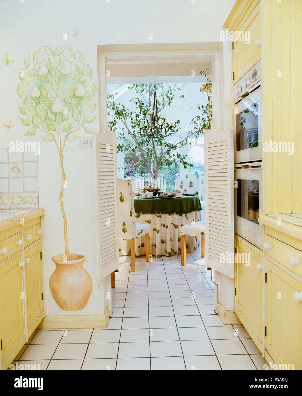 Stenciled Baum im Topf auf Wand der Küche mit hellgelben Einheiten und der Speisesaal durch offene Tür Stockfoto