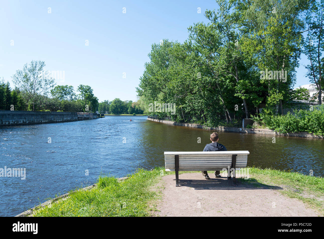 Ein junger Mann sitzt auf einer Bank in der Nähe des Flusses und sieht im Sport Boot. Stockfoto