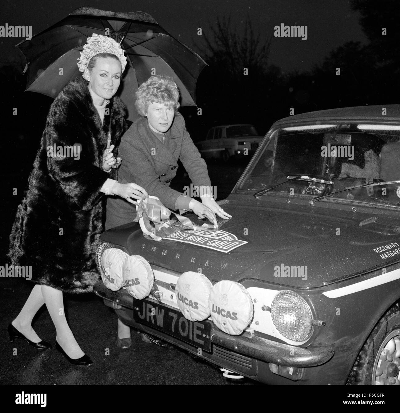 Dublin - Rosemary Smith (l) und ihre Beifahrer, Margaret Lowry, von Winslow, Buckinghamshire, Unterschlupf unter einem Regenschirm, wie sie aus dem Abdeckband die Rallye Monte Carlo Nummer auf Ihrem Sunbeam Imp Auto im Hyde Park, London, geboren. Stockfoto
