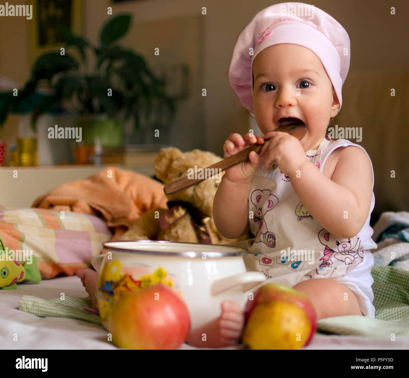 Baby Mädchen gekleidet wie ein Koch mit der Kappe ein Chef auf dem Kopf, hält eine hölzerne Löffel in einem Kochtopf und ein paar Äpfel mit einige Spielzeuge im Hinterg Stockfoto