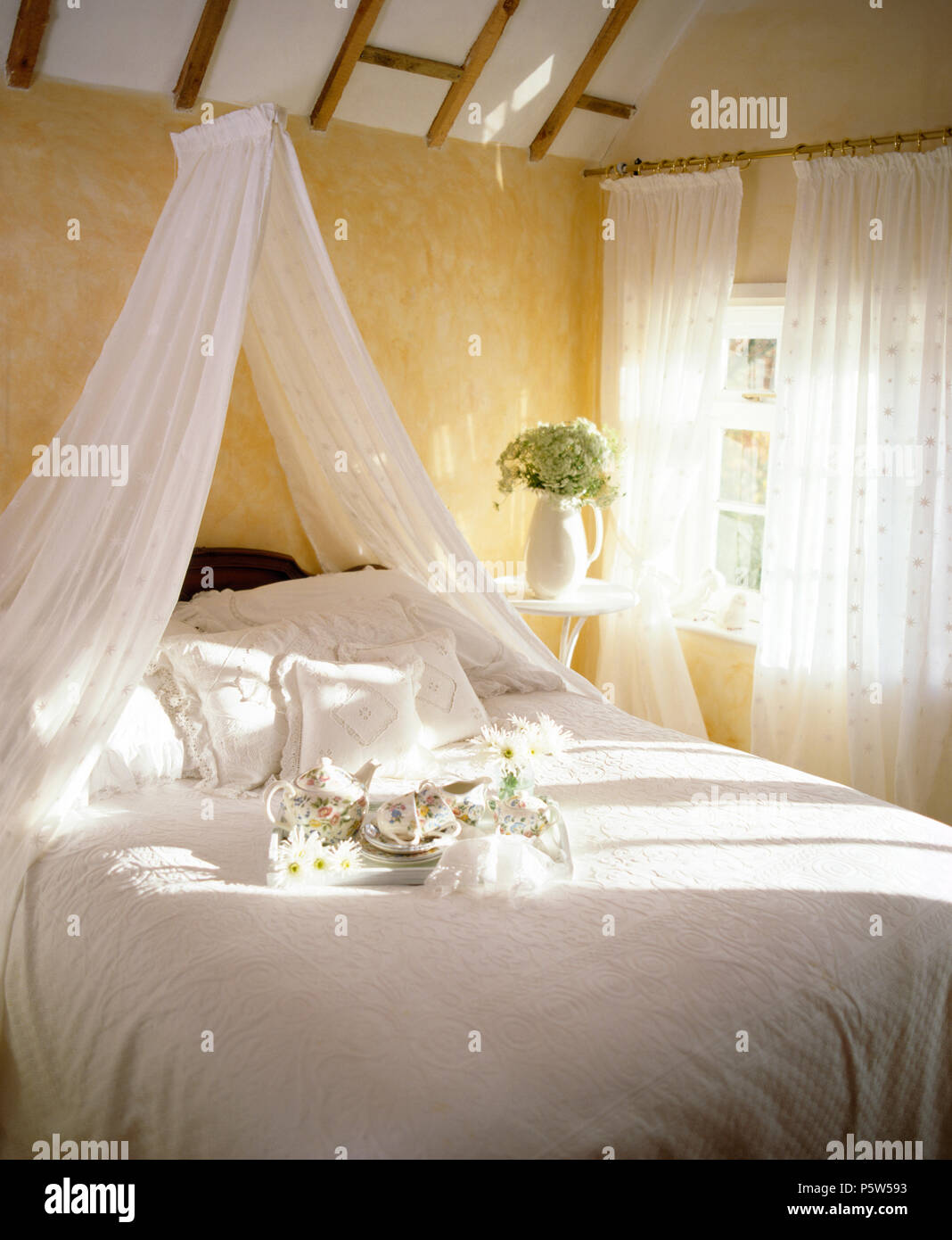 Weißer voile Gardinen über dem Bett mit weißer Bettwäsche im Cottage Schlafzimmer mit weißem voile Gardinen am Fenster Stockfoto