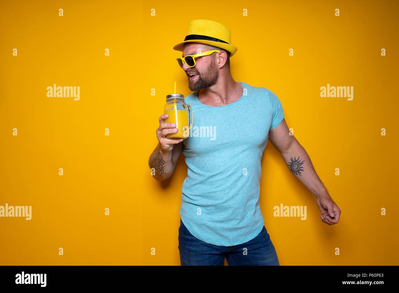 Kreative Mann in Sonnenbrille und Sommer Kleidung in gelben und blauen Farben stehend mit der Hand berühren stylischen Hut und Trinken von Creative cocktail Flasche entspannt auf Gelb studio Hintergrund. Stockfoto