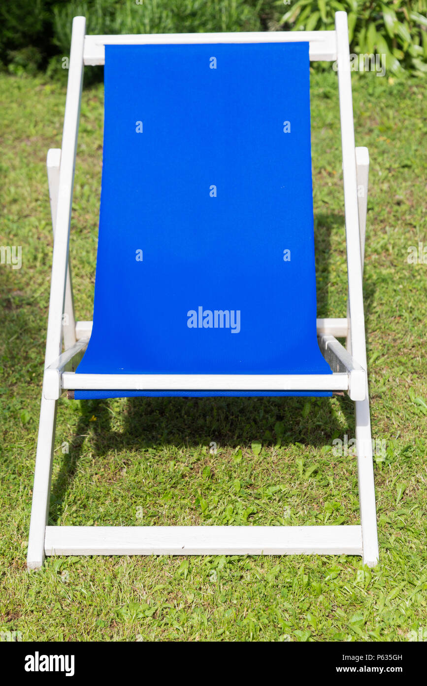 Nahaufnahme eines weißen und blauen Liegestuhl. Konzept der Entspannung während der Feiertage über eine grüne Wiese mit dem Hintergrund der Blumen und Pflanzen. Stockfoto