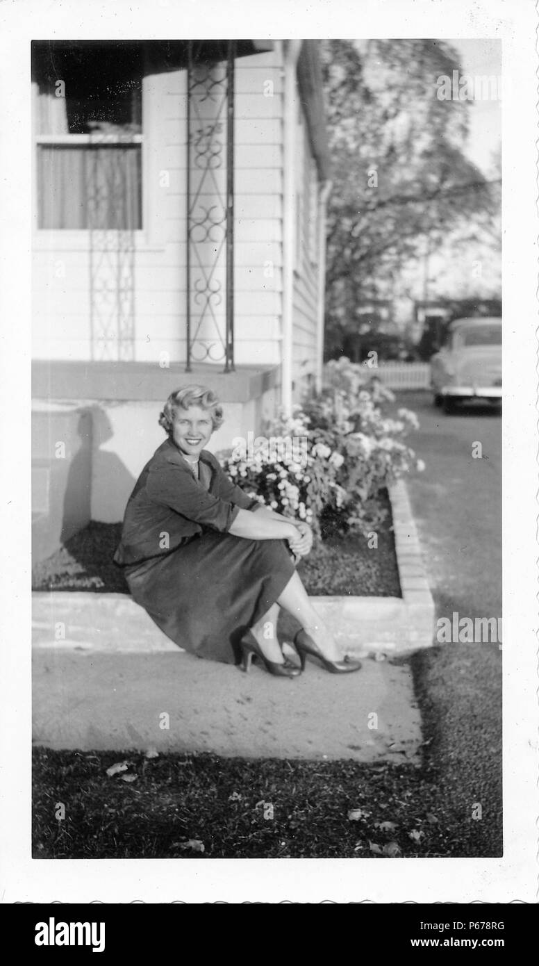 Schwarz-weiß Foto, zeigt eine attraktive, lächelnde Frau mit kurzen, lockige, blonde Haare, trug einen Rock und Highheels, und sitzen auf die konkrete Grenze ein Blumenbeet vor einem kleinen Haus, mit einem Oldtimer im Hintergrund sichtbar, wahrscheinlich in Ohio in den zehn Jahren fotografierte nach dem Zweiten Weltkrieg, 1950. () Stockfoto