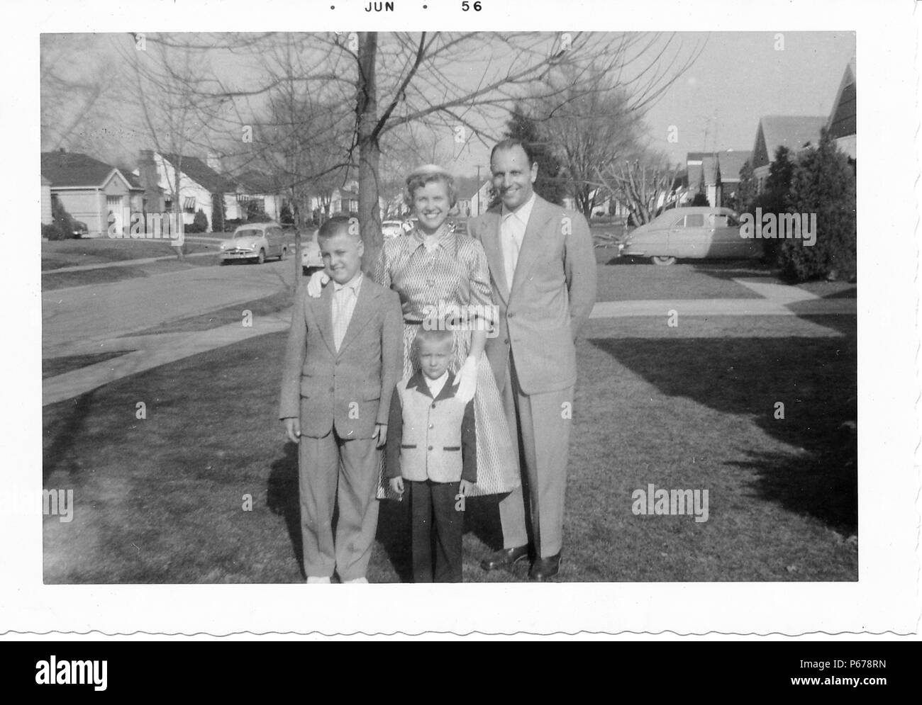 Schwarz-weiß Foto, zeigt eine Familie, in der vollen Länge, zusammen Posieren im Freien, der Mann mittleren Alters, mit einem zurücktretenhaarstrich, trägt einen hellen Anzug und steht mit einem kurzhaarigen Frau, trug einen Rock, Bluse und Handschuhe, und zwei Jungen, beide trugen Anzüge, die Kleinere trägt eine zweifarbige Jacke, mit Bäumen, Oldtimer und s-Häuser im Hintergrund sichtbar, wahrscheinlich in Ohio, Juni, 1956 fotografiert. () Stockfoto