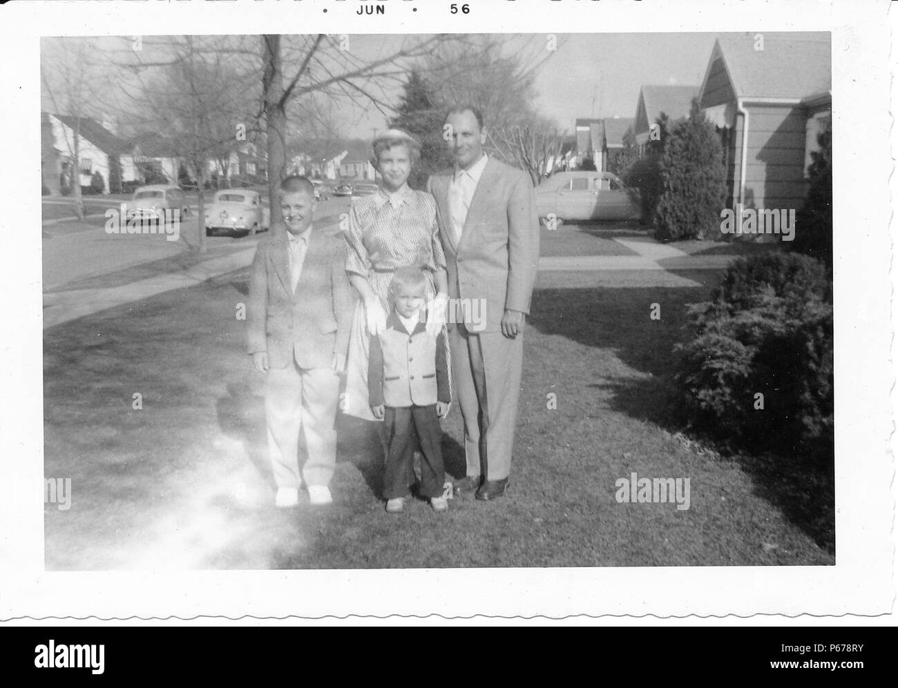 Schwarz-weiß Foto, zeigt eine Familie, in der vollen Länge, zusammen Posieren im Freien, der Mann mittleren Alters, mit einem zurücktretenhaarstrich, trägt einen hellen Anzug und steht mit einem kurzhaarigen Frau, trug einen Rock, Bluse und Handschuhe, und zwei Jungen, beide trugen Anzüge, die Kleinere trägt eine zweifarbige Jacke, mit Bäumen, Oldtimer und s-Häuser im Hintergrund sichtbar, wahrscheinlich in Ohio, Juni, 1956 fotografiert. () Stockfoto