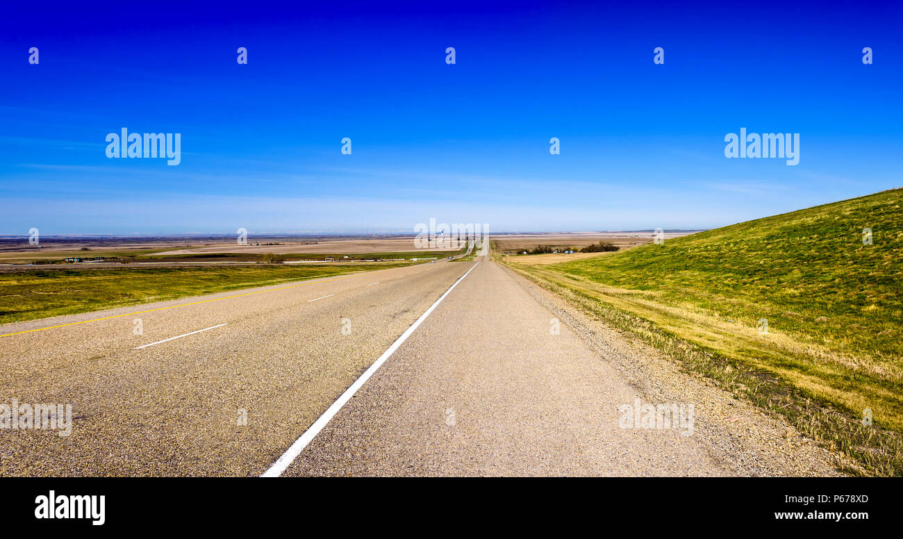 Panorama über eine lange Asphaltstraße zwischen Feldern mit grünem Gras, einem dichten blauen Himmel im Sommer Stockfoto