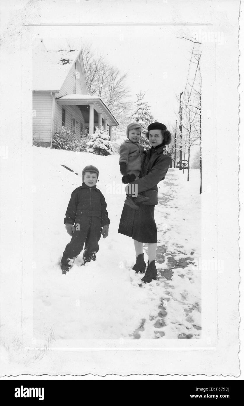 Schwarz-weiß Foto, zeigt eine dunkelhaarige Frau, in voller Länge, trägt eine Baskenmütze, kurze Stiefel, Rock und Jacke, stehend auf einem verschneiten Gehweg, mit einer kleinen, gemütlich Kind in ihren Armen gekleidet, mit einem zweiten Junge, trägt ein Schneeanzug, Mütze, stand neben ihr, mit schneebedeckten Bäumen und einem Abstellgleis bedeckt im Hintergrund, wahrscheinlich in Ohio in den zehn Jahren fotografierte nach dem Zweiten Weltkrieg, 1950. () Stockfoto