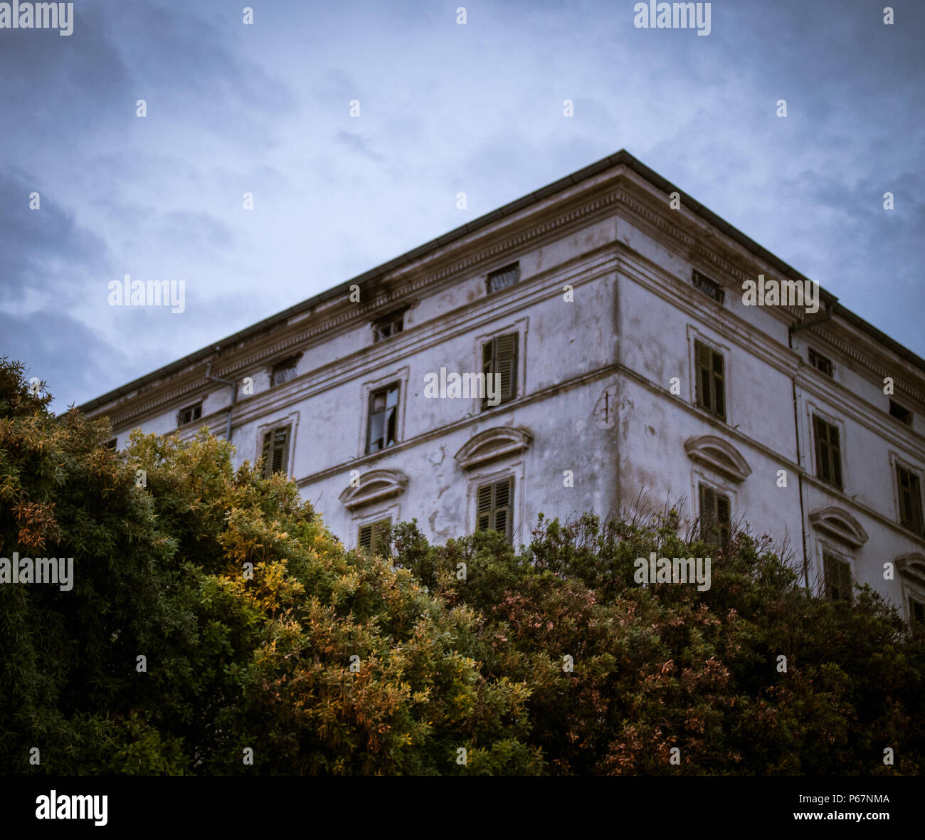 Die Ecke eines alten europäischen haunted House / Villa im Stil mit kaputten Fenstern von Bäumen unter einer Dämmerung Himmel umgeben verloren. Stockfoto