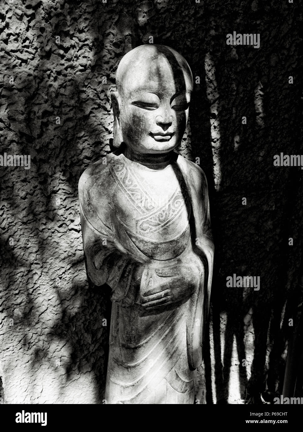 Eine schwarz-weiße Studie einer Kannon Bodhisattva-Statue aus Stein, aufgenommen mit dramatischen kontrastreichen Schatten vor einer strukturierten Steinkulisse. Stockfoto