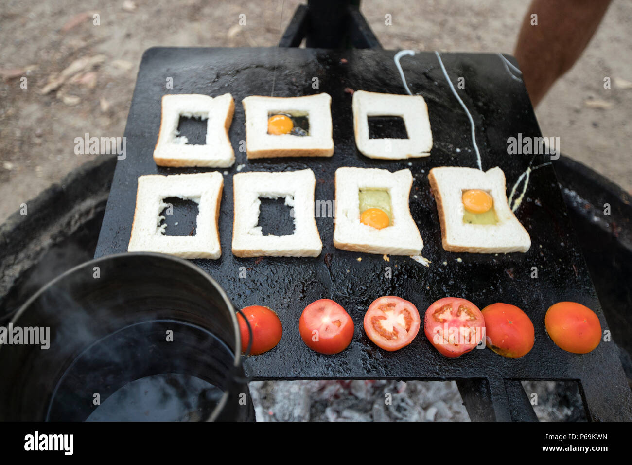Scheiben Toast mit einem ausgestochener Mitte liegen auf der Grillplatte über dem Feuer, einige bereits mit Ei gefüllt, halbierte Tomaten daneben aufgereiht, ein Frühstück im Outback-Stil im Kakadu-Nationalpark, Northern Territory, Australien. Stockfoto