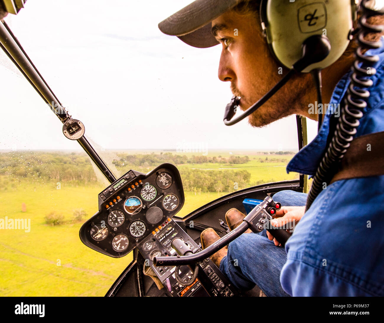 Pilot Jock konzentriert sich auf den Steuerknüppel seines Robinson R44-Hubschraubers, während üppige grüne Weiden in der Nähe von Rakula, Northern Territory, Australien, unter ihm vorbeiziehen. Stockfoto