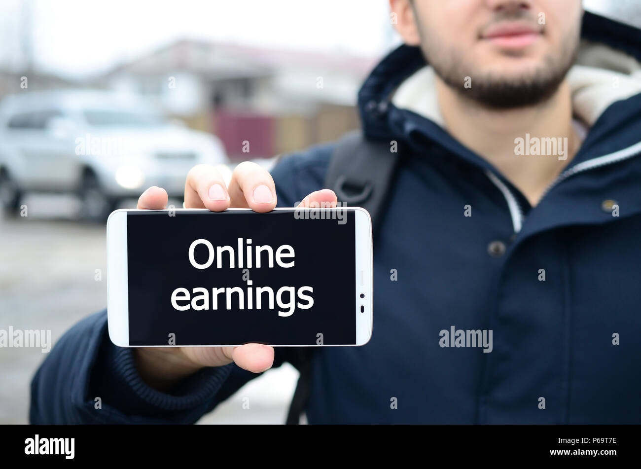 Ein junger Kerl zeigt eine Inschrift auf dem Smartphone Display auf dem Hintergrund eines Autos und ein großes Haus im Winter. Online Ergebnis. Stockfoto