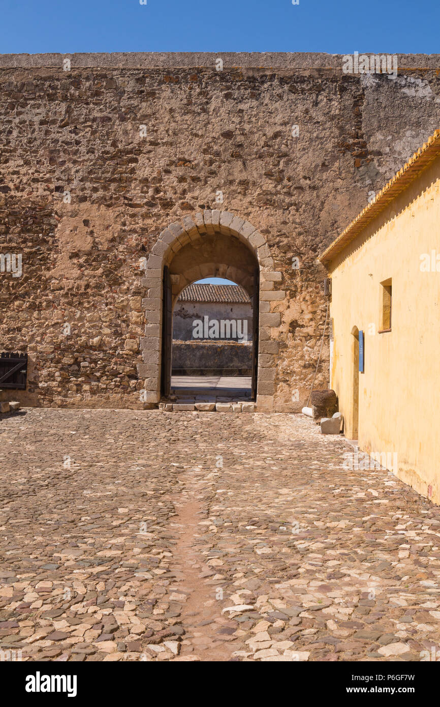 Kopfsteinpflaster Platz mit einem gelben Haus und Festung aus Stein mit einem Tor mit einem Bogen. Strahlend blauen Himmel. Castro Marim, Algarve, Portugal. Stockfoto