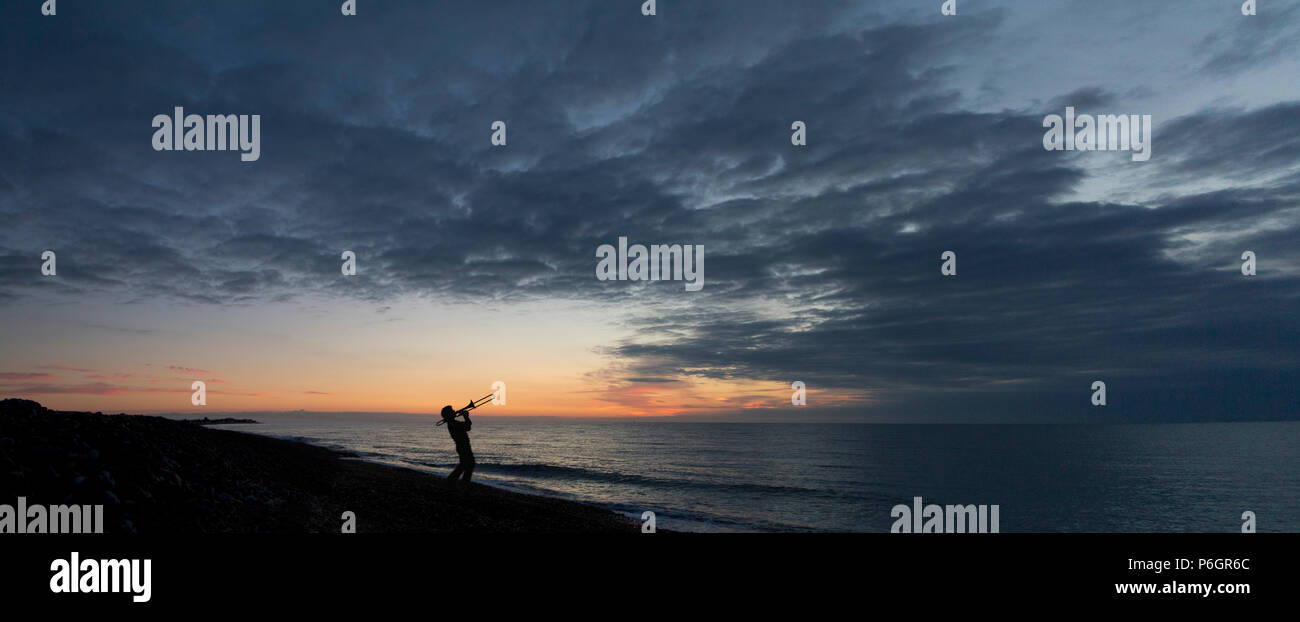 Panorama von einem Mann in Silhouette spielen eine Posaune gegen einen Sonnenaufgang am Strand mit frühen Morgen Wolken Stockfoto