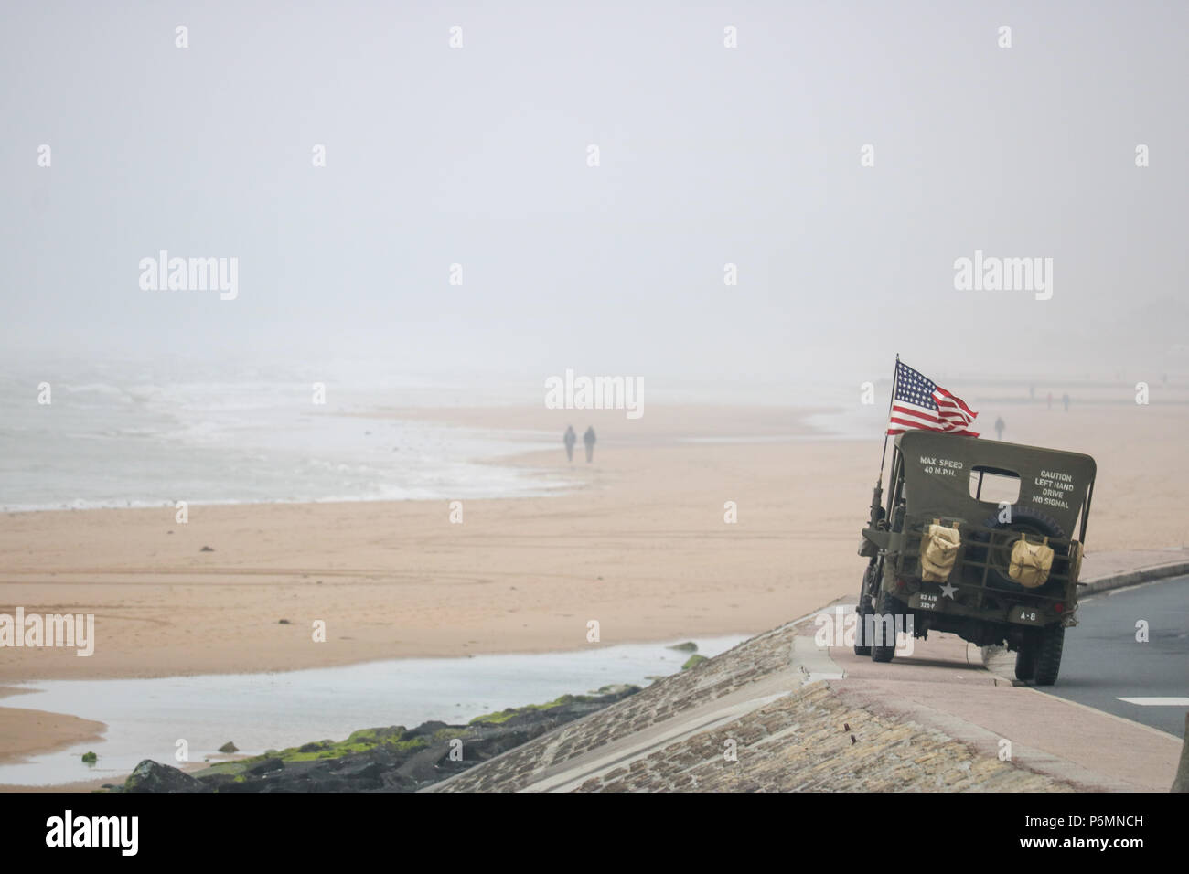 Vierville-sur-Mer, Frankreich. Ein Willys Jeep fliegen die US-Flagge sitzt auf der Ufermauer mit Blick auf Omaha Beach, als entfernte Paar Spaziergang entlang der Waters Edge am Jahrestag des D-Day Stockfoto