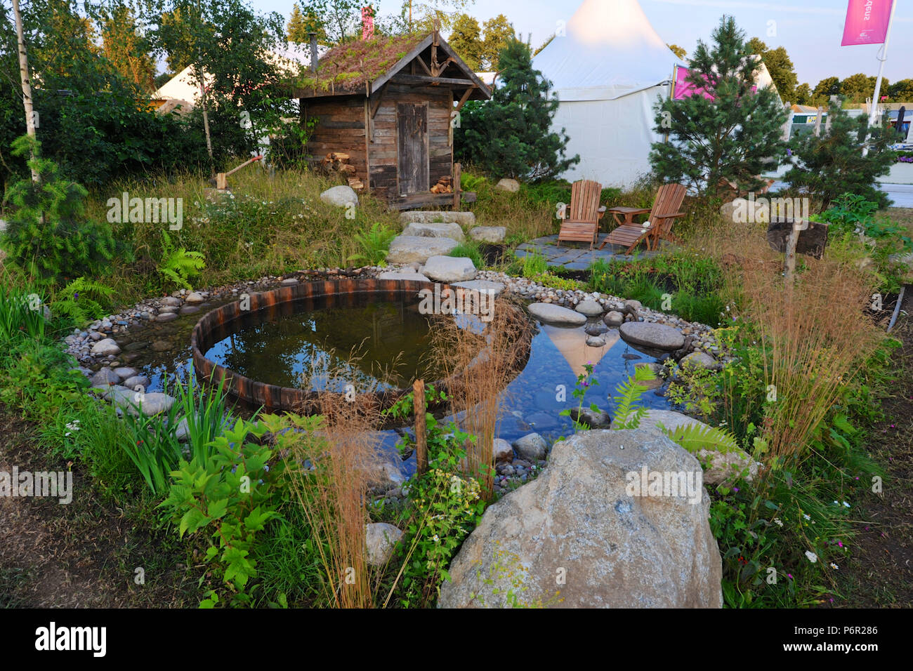 London, Großbritannien. 2. Juli 2018. Die Viking Kreuzfahrten Nordic Lifestyle Garten (von Stephen Hall) Eine der schönen und eleganten Gärten zeigen, die auf der 2018 RHS Hampton Court Flower Show, die heute auf dem Gelände der Hampton Court Palace, London, Großbritannien, 02. Juli 2018 eröffnet. Neuerstellen eines Nordischen Waldgebiet führt ein Pfad zu Stein Sitzbereich neben der traditionellen Sauna Hütte gepflastert. Nordic Stein Geröll führen dann zu einem Tauchbecken, gesäumt mit Kopfsteinpflaster und kleinen Felsbrocken. Größere Felsbrocken scheinen durch Bereiche der wiese gras rund um die Sauna wächst, während die Nordischen Bäume und Sträucher Stockfoto