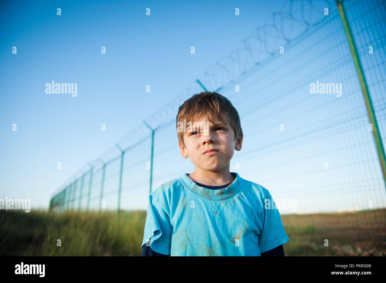 Süße nachdenkliche schmutzige Gesicht Kind Flüchtling Portrait auf Zaun mit Stacheldraht bakdrop im Sonnenuntergang Stockfoto