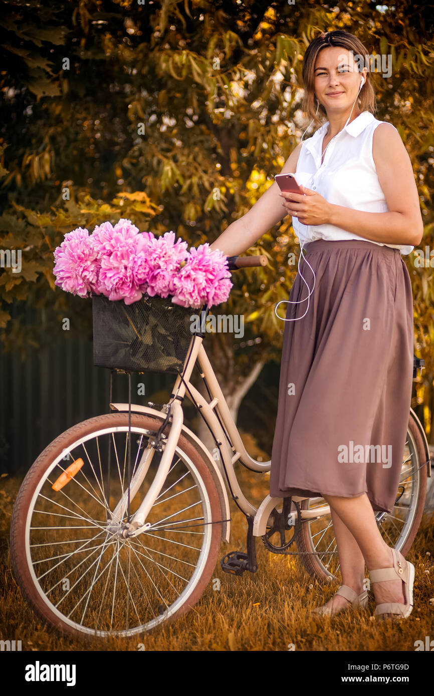 Junge dunkelhaarige Frau in einem weißen Hemd beige Rock hört Musik auf dem Telefon, und steht mit einem Fahrrad mit einem Metallkorb mit einem großen bouqu Stockfoto