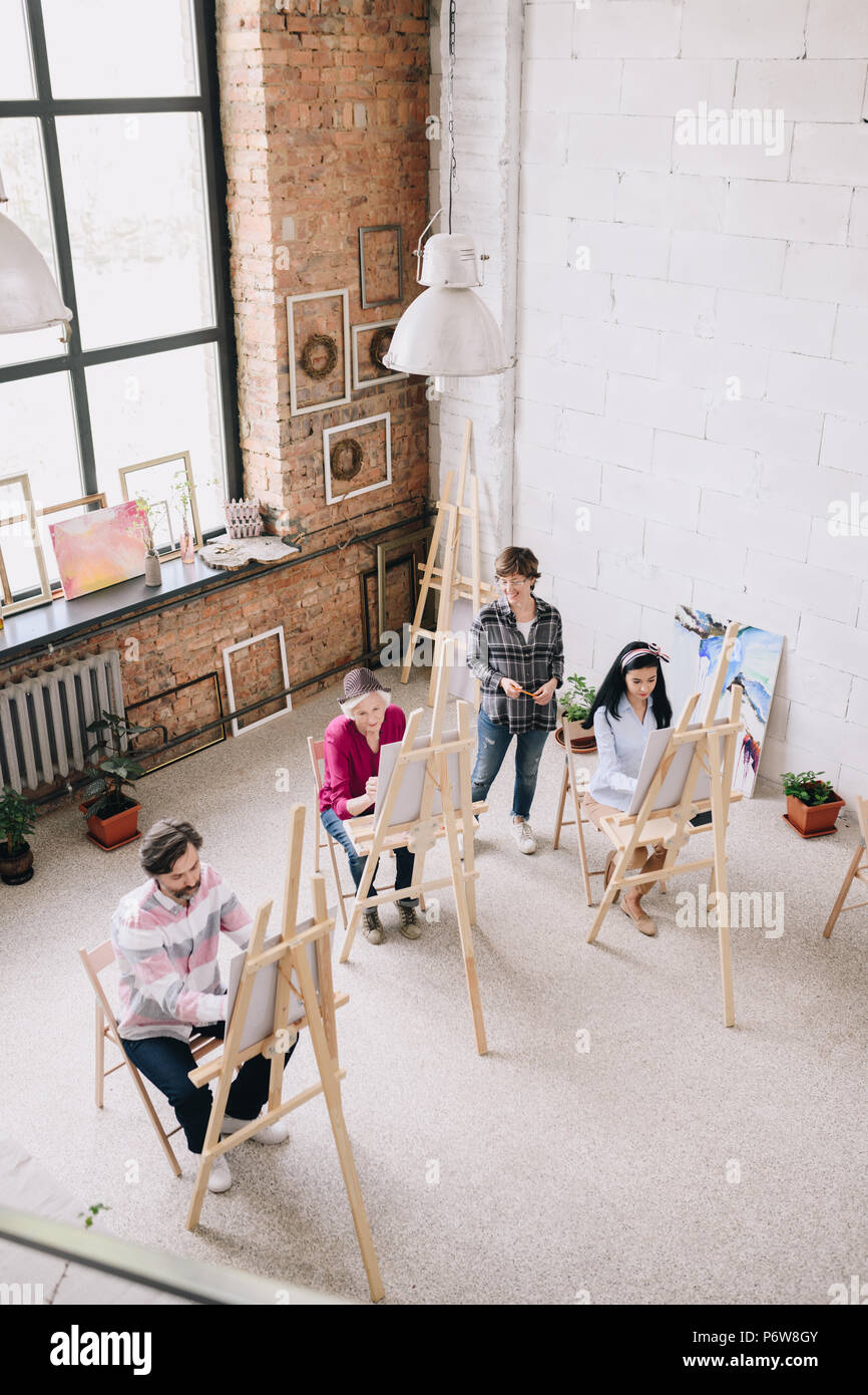 Oben Ansicht Portrait von erwachsenen Kursteilnehmer Malerei sitzt von staffeleien in art studio mit weiblichen kunst Lehrer beobachten Sie in geräumigen und lichtdurchfluteten Dachboden, c Stockfoto