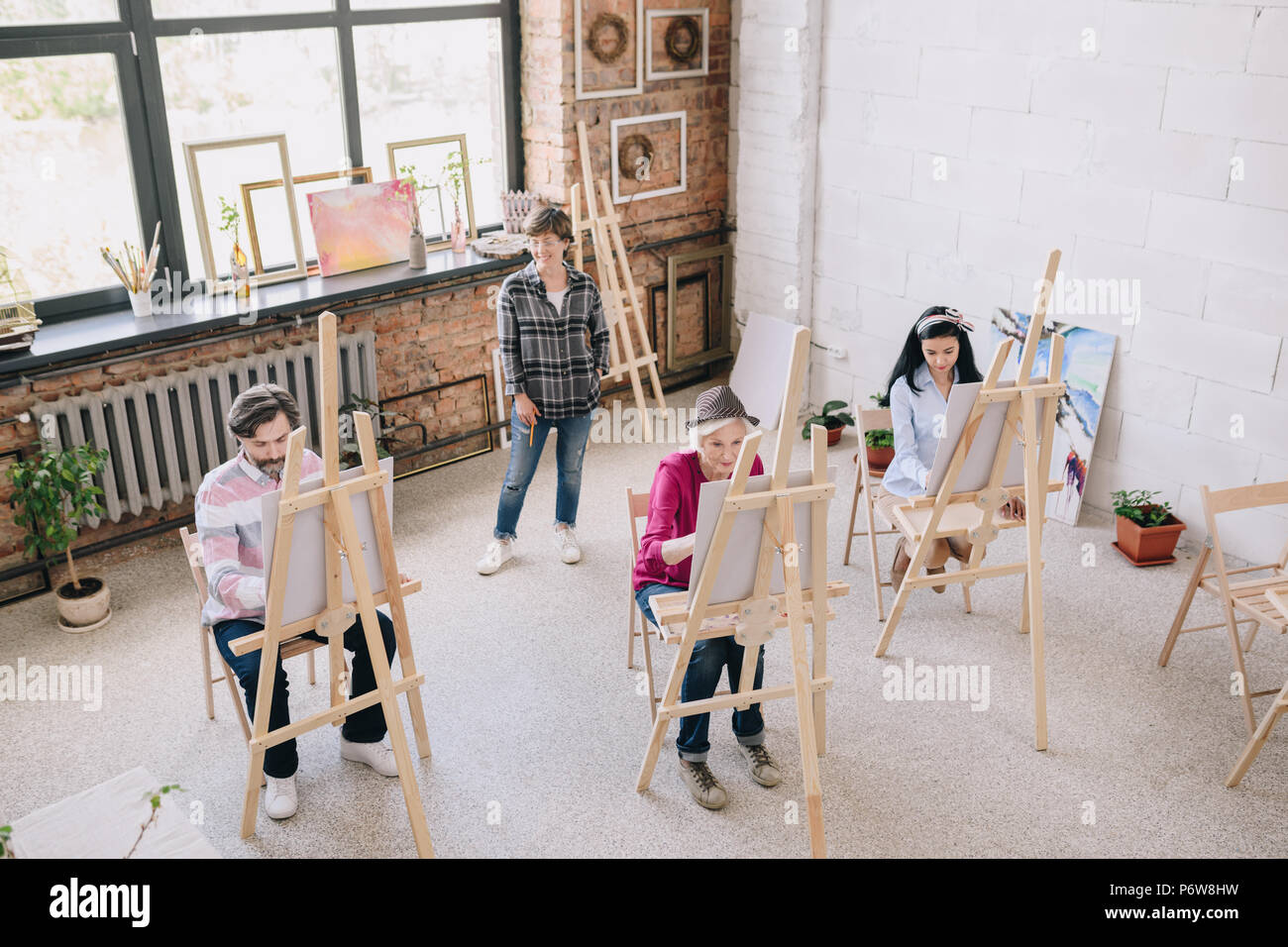 Hohen winkel Portrait von erwachsenen Kursteilnehmer Malerei sitzt von staffeleien in art studio mit weiblichen kunst Lehrer beobachten Sie in geräumigen und lichtdurchfluteten Dachboden, c Stockfoto