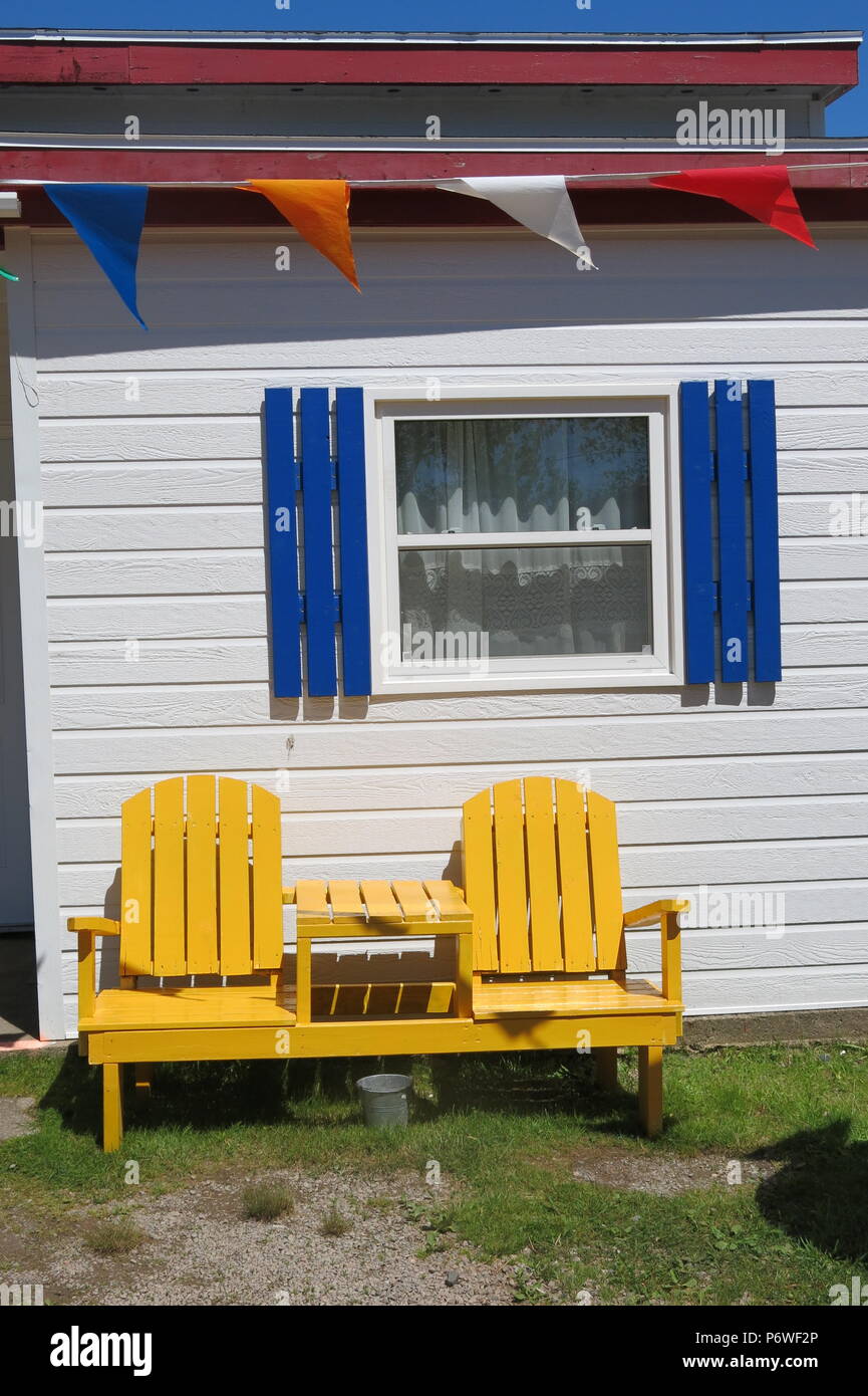 Zwei Sitzbank und Tisch im Adirondack Stil, in einem hellen Gelb, gegen ein weißes Haus mit blauen Fensterläden weatherboard Stockfoto