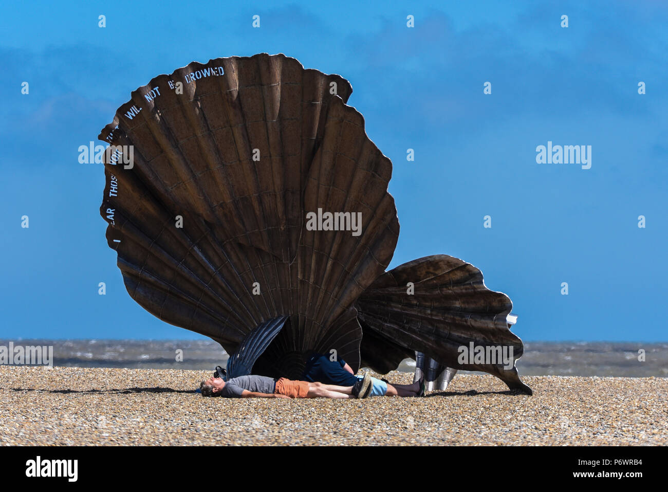 Menschen, die Zuflucht von den schweren blustery Wind hinter der Jakobsmuschel Skulptur auf Henne Strand. Obwohl Sunny ein starker Wind wehte in der Nordsee. Die Skulptur namens Jakobsmuschel, Benjamin Britten, der am Strand entlang an den Nachmittagen zu gehen. Aus Edelstahl von Suffolk erstellt Künstler Maggi Hambling Stockfoto