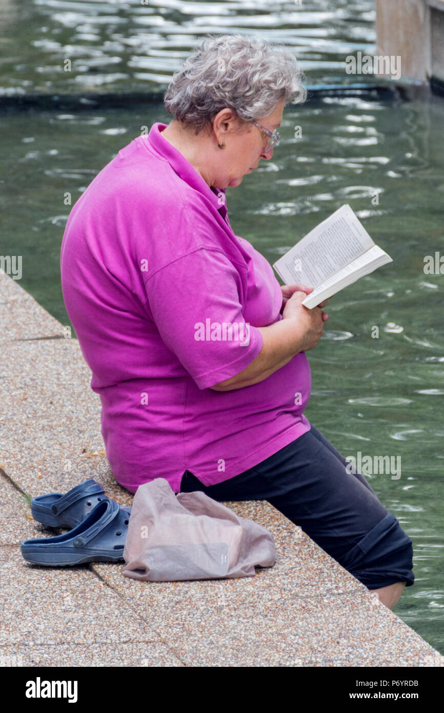 Niska Banja, Serbien - Juli 02, 2018: Alte Frau mit Buch mit Beinen in gesundes Wasser im Kurpark. Urlaub und Genuss des Sommers Konzept Stockfoto