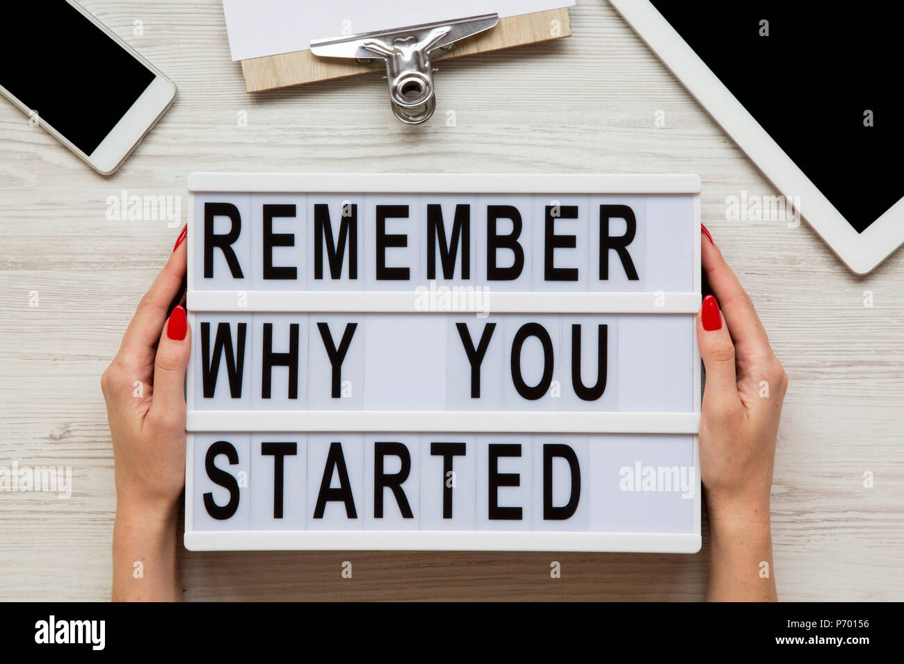 Arbeitsbereich mit Smartphone, Tablet, Blatt und "warum Sie gestartet' Worten auf Lightbox auf weißem Schreibtisch aus Holz erinnern, Ansicht von oben. Weibliche Hände halten moderne Bo Stockfoto