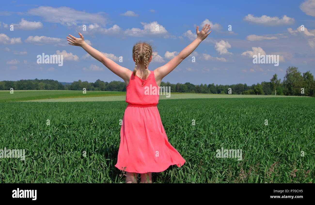 Junge Frau, blonde Haare, kurze Zöpfe, steht auf dem Rücken im grünen Feld, auf dem Land, Sommer, im rosa Kleid mit offenen Rams gekleidet, Freuden Stockfoto
