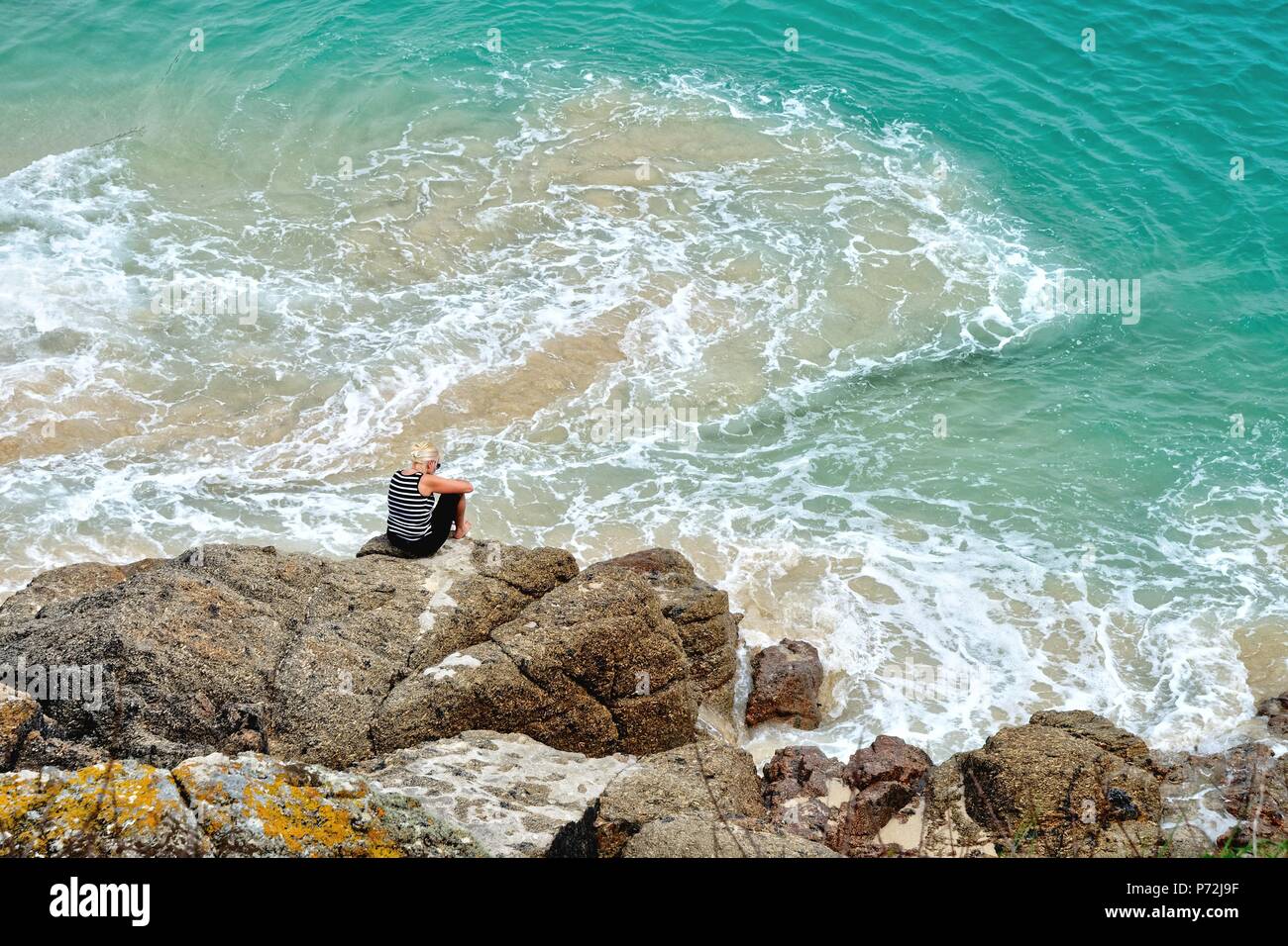 Im mittleren Alter blonde Frau, sitzen auf den Felsen durch einen Cornish blue sea Porthcurno Cornwall England UK umgeben Stockfoto