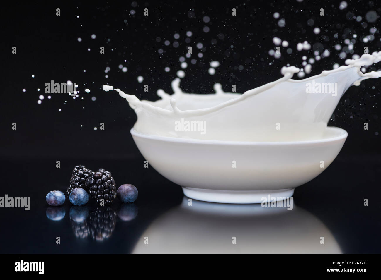 Saftige Beeren durch weiße Schüssel mit Milch spritzen auf schwarzem Hintergrund Stockfoto