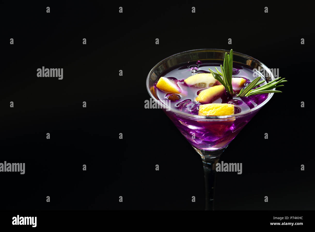 Nahaufnahme eines frischen Sommer Purple Rain Cocktail in einem Martini Glas mit Eis, Zitronenscheiben und Thymian auf eine schwarze texturierte Oberfläche Stockfoto