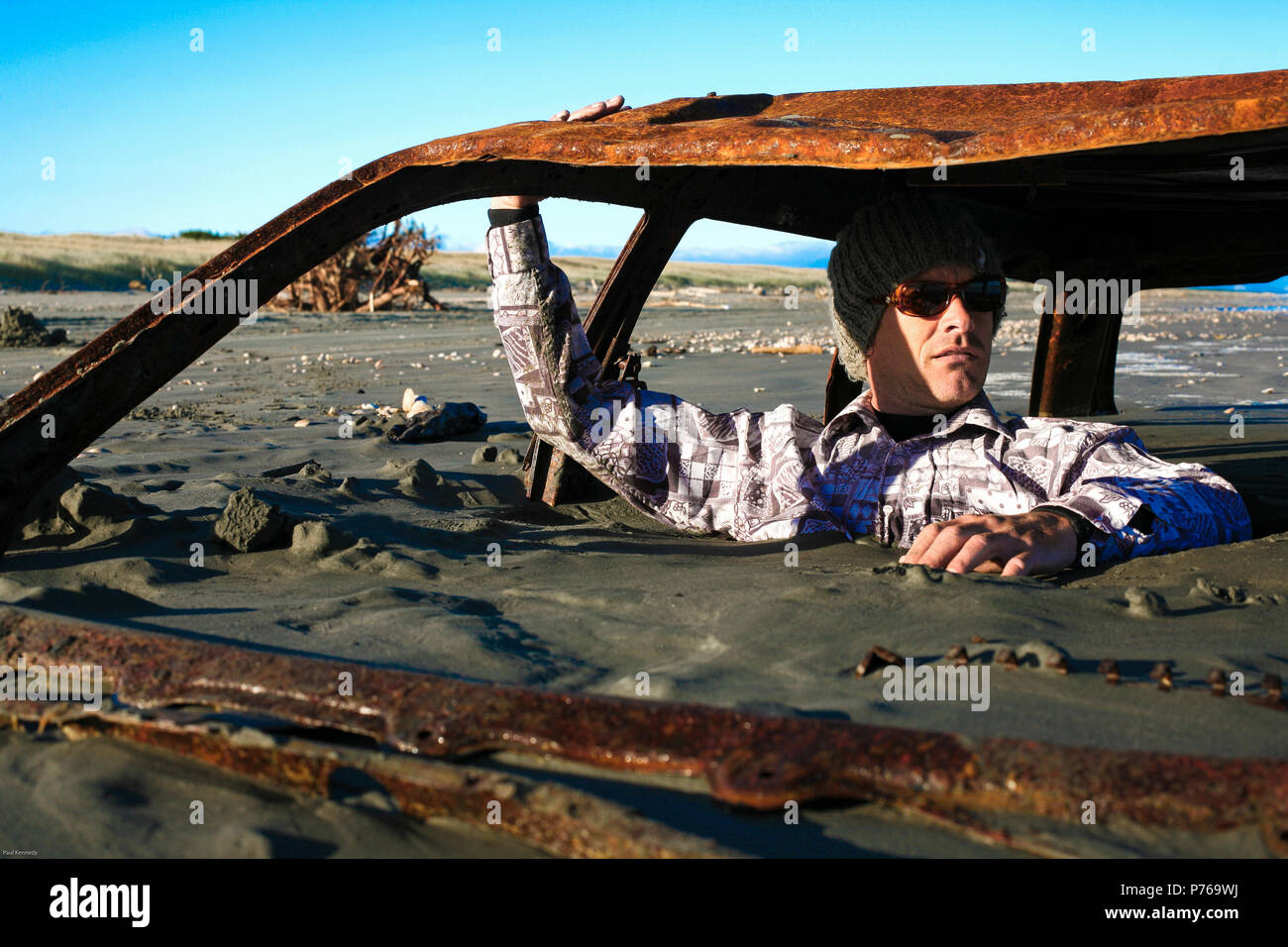 Mann sitzt auf verrosteten Autowrack vergraben im Sand am Strand Stockfoto