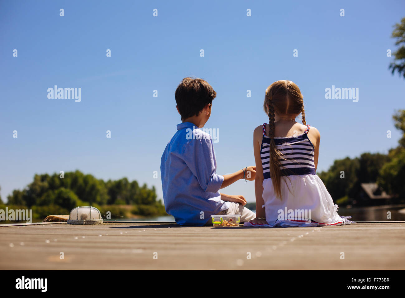 Süße kleine Geschwister sitzen auf dem Pier und Snacks in Stockfoto