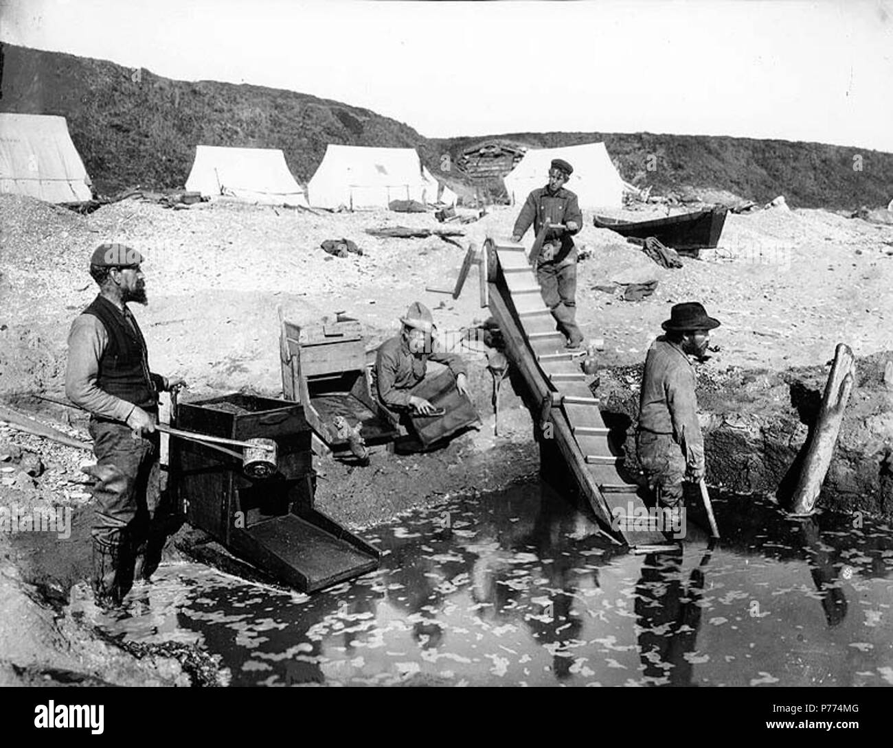 . Englisch: Bergbau Betrieb auf Nome, Alaska, Ca. 1900. Englisch: Drei Männer und eine Frau, die Rocker in den Sand mit Zelten in den Hintergrund. Bildunterschrift auf das Bild: "Rocking auf Nome Strand." Original Foto von Eric A. Hegg 1281 A; kopiert von Webster und Stevens 337. a. Nome Gold Rush. Themen (LCTGM): Gold Miner - Alaska - Nome; Gold mining - Alaska - Nome; Strände - Alaska - Nome; Bergbau Camps - - Alaska - Nome Themen (LCSH): Alaska - Gold Entdeckungen. ca. 1900 9 Bergbau Betrieb auf Nome, Alaska, ca 1900 HEGG (5) Stockfoto