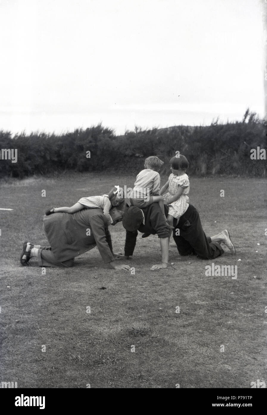 1950, Spaß clmbing auf ihrem Vati zurück..... historische Bild von drei kleinen Kindern liegen und Reiten auf dem Rücken von ihren Vätern, die auf Ihre Hände und Knie auf dem Gras außerhalb, England, UK. Stockfoto
