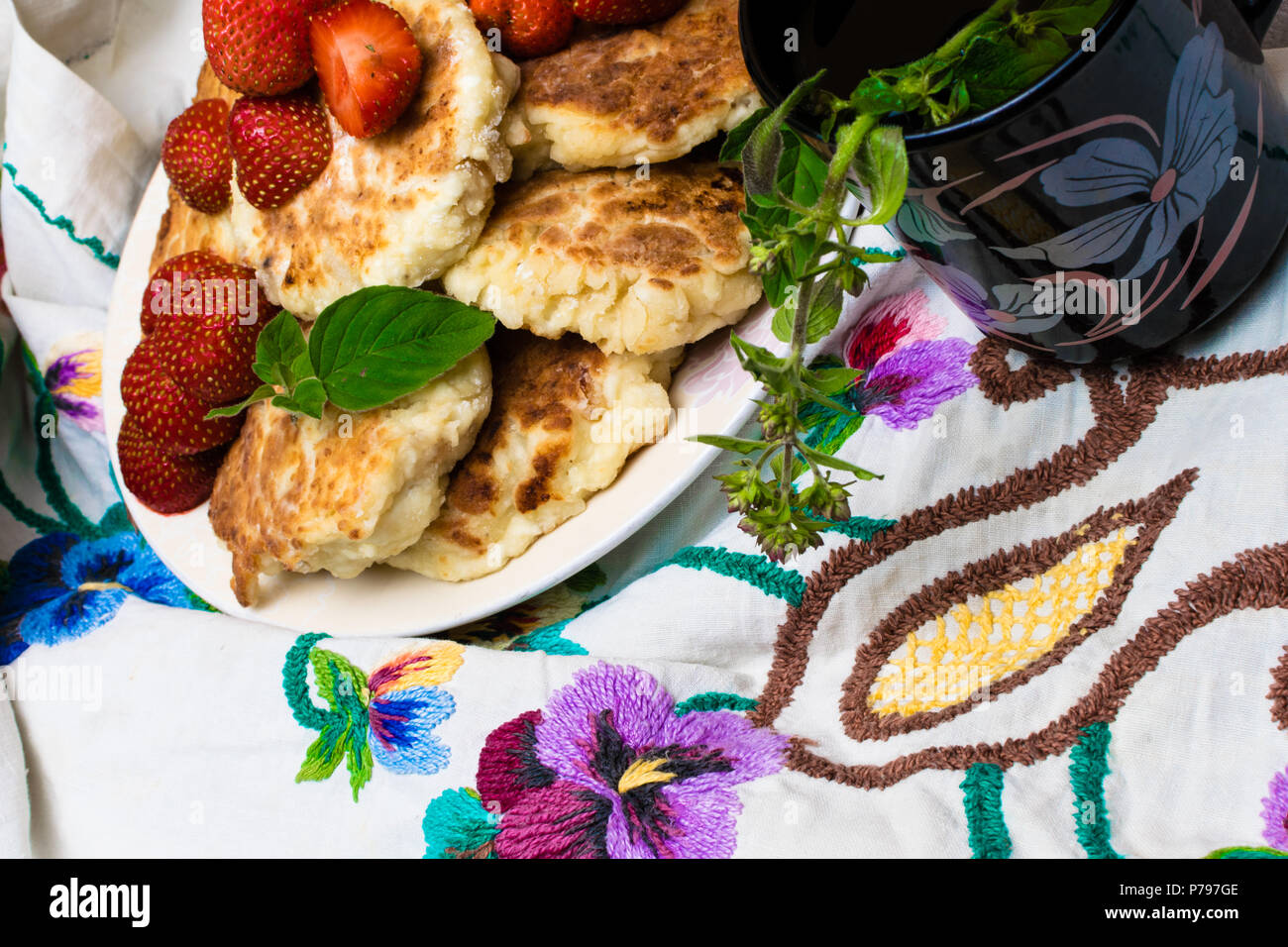 Frisch gebackene Quark Pfannkuchen mit Rosinen in einem weissen Teller, Tasse Tee mit Minze, Melisse Zweig, bestickt mit Farbe Blumen Tischdecke. Zu Stockfoto