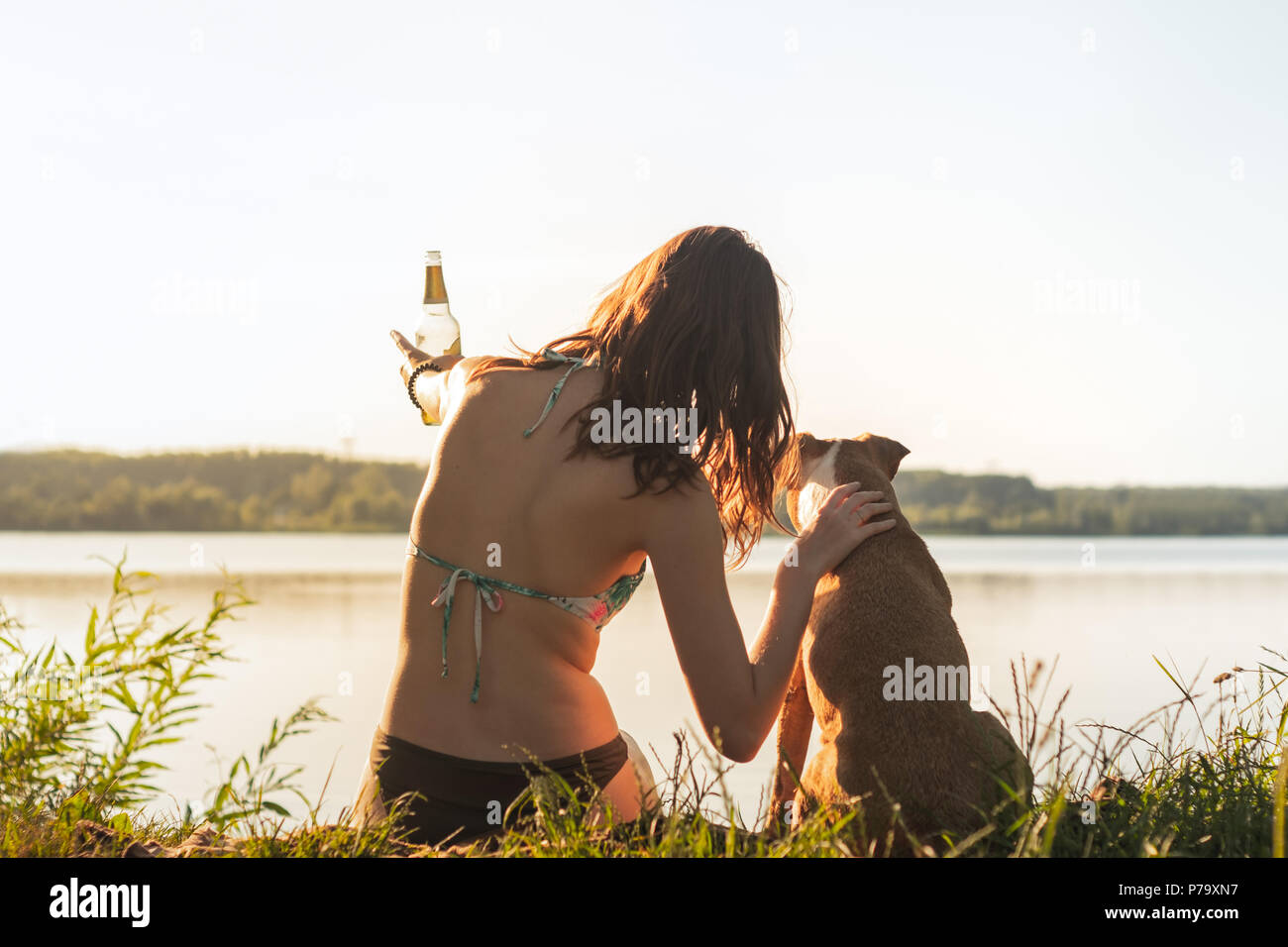 Schöne schlanke Frau mit Hund genießen Sie einen wunderschönen Blick in der Nähe des Sees bei Sonnenuntergang von warmen und sonnigen Nachmittag. Junge Passform weiblichen in einem alkoholfreien Getränk am Fluss Stockfoto