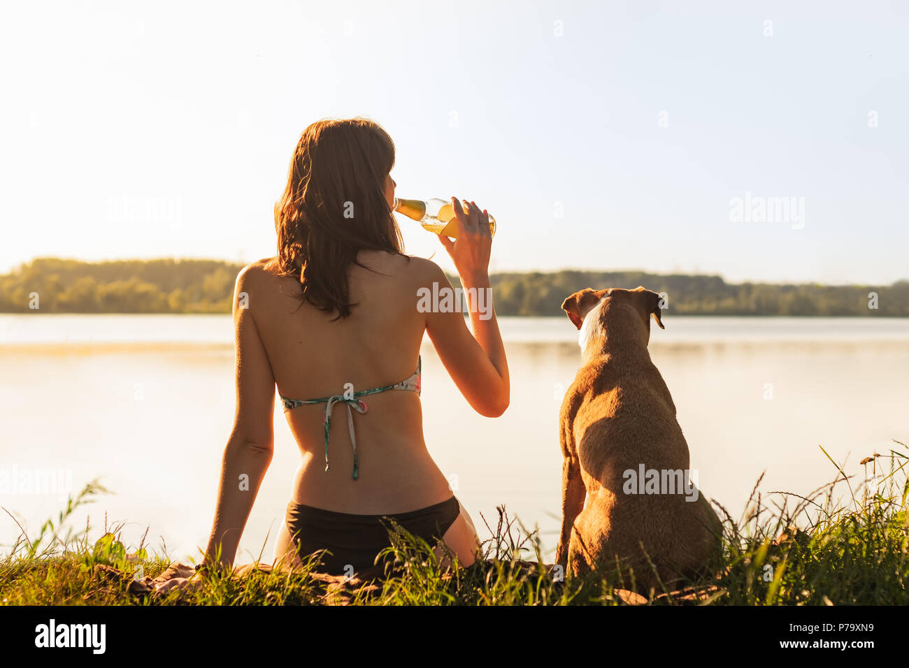 Schöne schlanke Frau mit Hund genießen Sie einen wunderschönen Blick in der Nähe des Sees bei Sonnenuntergang von warmen, sonnigen Nachmittag und einem Drink. Junge Passform weiblichen Sitzen wi Stockfoto