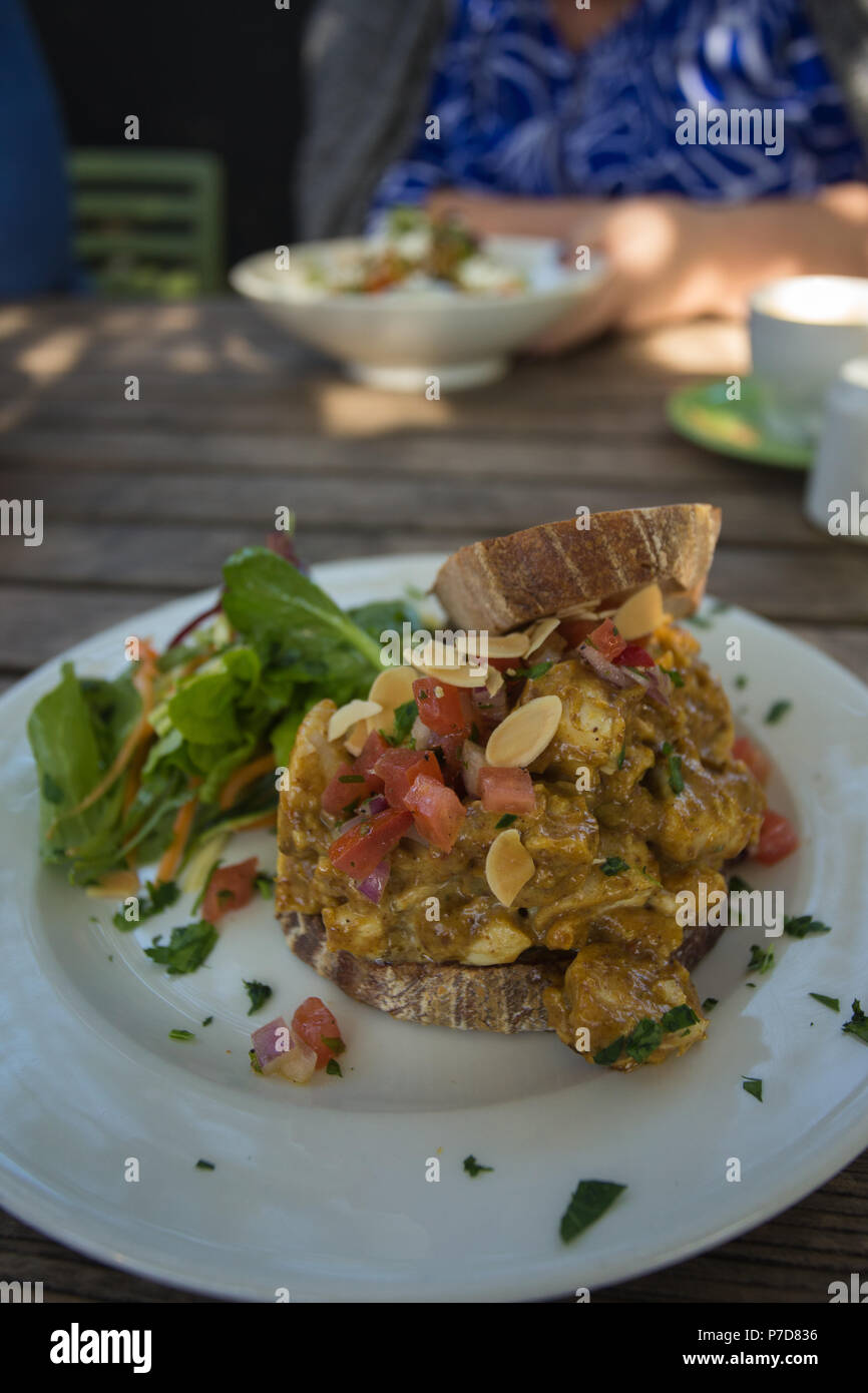 Offenes Sandwich mit gegrilltem Hähnchen in würzigem Harissa und Dattendressing auf weißem Teller auf einem Holzrosttisch mit Person im Hintergrund Stockfoto