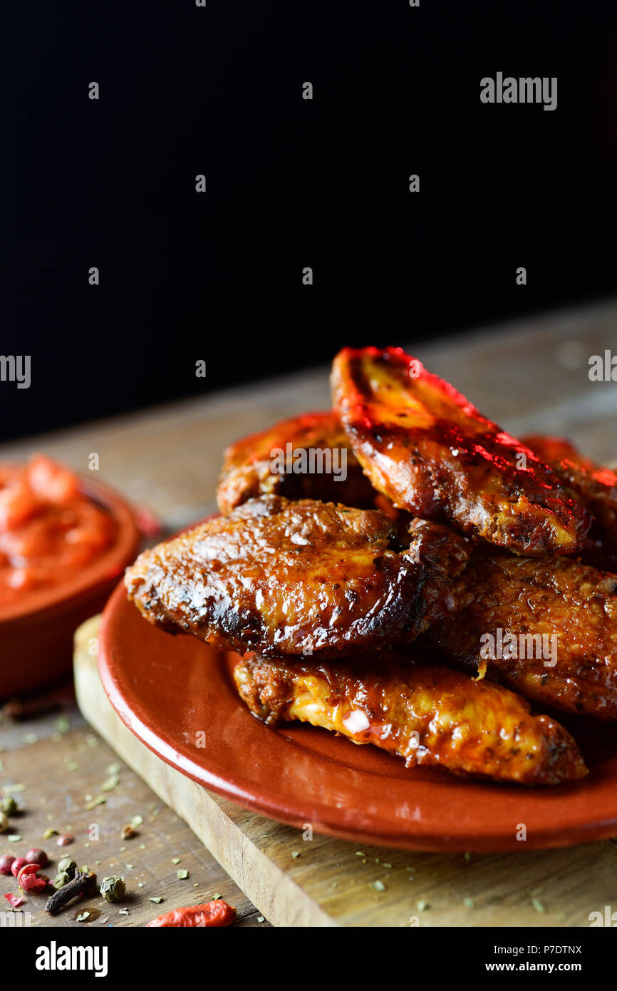Nahaufnahme von einigen barbecue Chicken Wings in einem Steingut Teller und eine Schüssel mit Barbecue Sauce auf einem rustikalen Holztisch, vor einem schwarzen Hintergrund wit Stockfoto