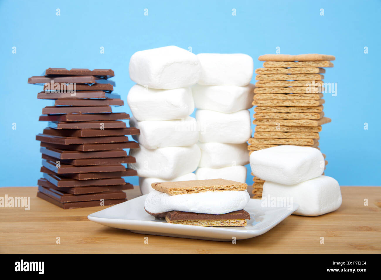 Hausgemachte s'More auf eine quadratische Platte mit Stapeln von graham Cracker, Schokolade und Marshmallows auf einem Holztisch mit hellblauem Hintergrund. Stockfoto
