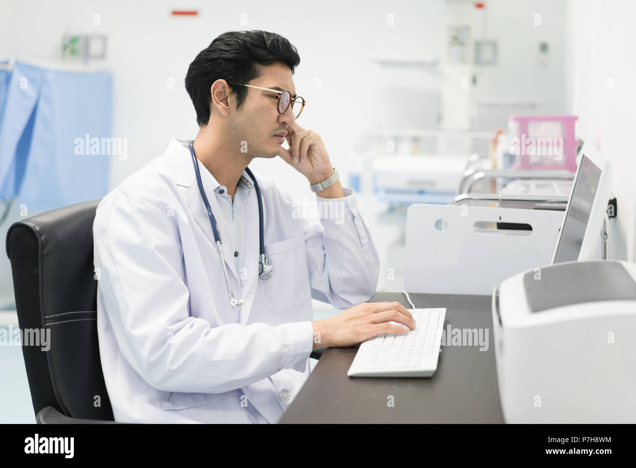 Portrait von Asiatischen schwere Arzt Mann mit einem Personal Computer im Krankenhaus. Gestörte Arzt am Schreibtisch sitzen, Sorgen, Denken schwer. Stockfoto