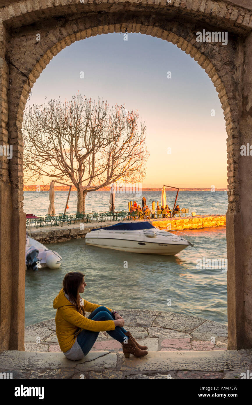Touristen bewundern Sie den Sonnenuntergang auf dem kleinen Hafen von Punta San Vigilio aus dem Bogen der charmante Inn aus dem 16. Jahrhundert am östlichen Ufer des Gardasees, in der Provinz Verona, Venetien, Italien. Stockfoto