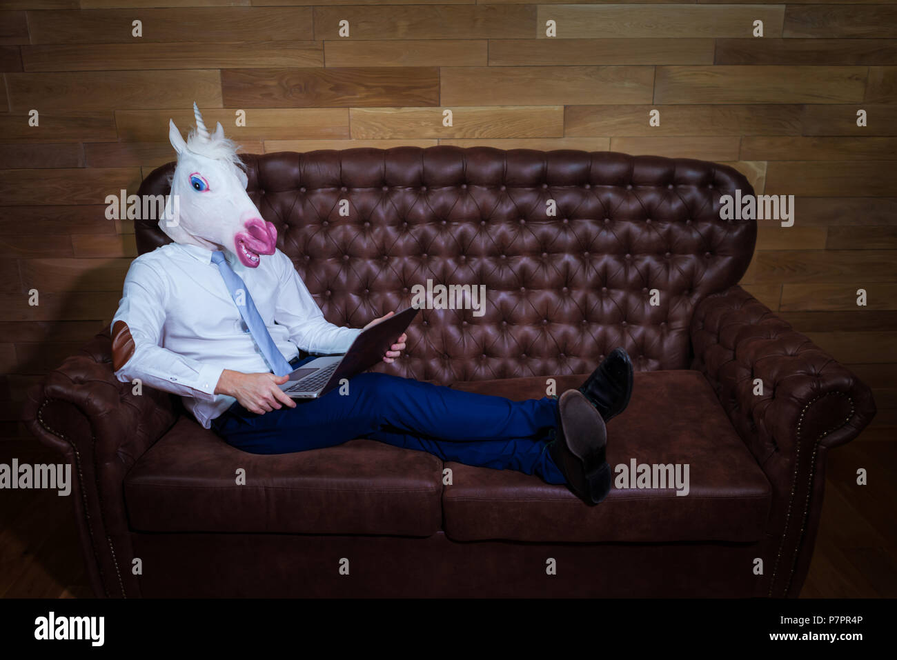 Ungewöhnliche Mann zu Hause arbeiten im Büro. Freaky junge Manager in komischen Maske auf dem Hintergrund der hölzernen Wand. Lustige Einhorn in Anzug sitzt auf einem Sofa wie ein Chef. Stockfoto