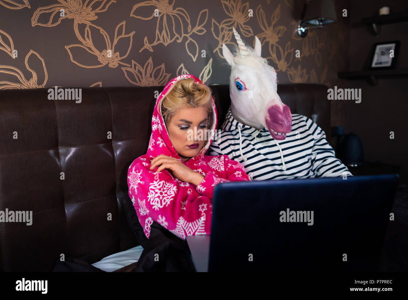 Schwere Mädchen liegt auf dem Bett mit seltsamen Freund in lustige Maske. Ungewöhnliche Paar schaut Film auf Laptop in der stilvollen Wohnung. Stockfoto