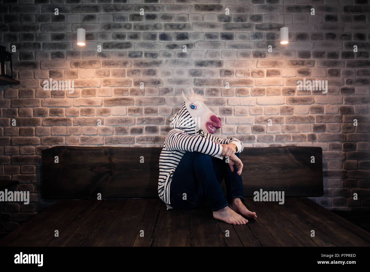 Ungewöhnliche junge Mann in komischen Maske sitzt alleine auf dem Bett in stilvolle Apartment. Traurige Einhorn in legere Kleidung. Freaky Mann nach oben verbringen Sie die Zeit zu Hause. Stockfoto