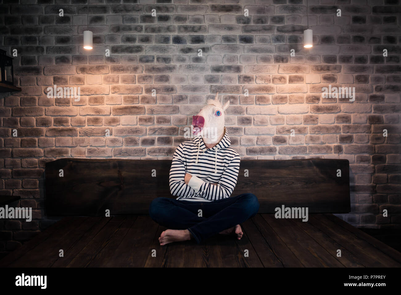 Ungewöhnliche junge Mann in komischen Maske sitzt alleine auf dem Bett in stilvolle Apartment. Ernsthafte Einhorn in legere Kleidung. Freaky Mann zu Hause. Stockfoto